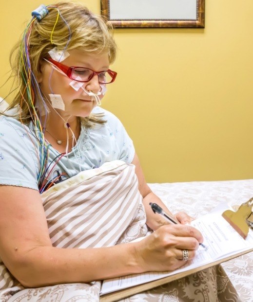Woman writing on clipboard while wearing sensors for sleep testing in Denton