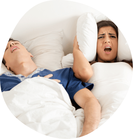 Angry woman in bed covering ears with pillow next to snoring man